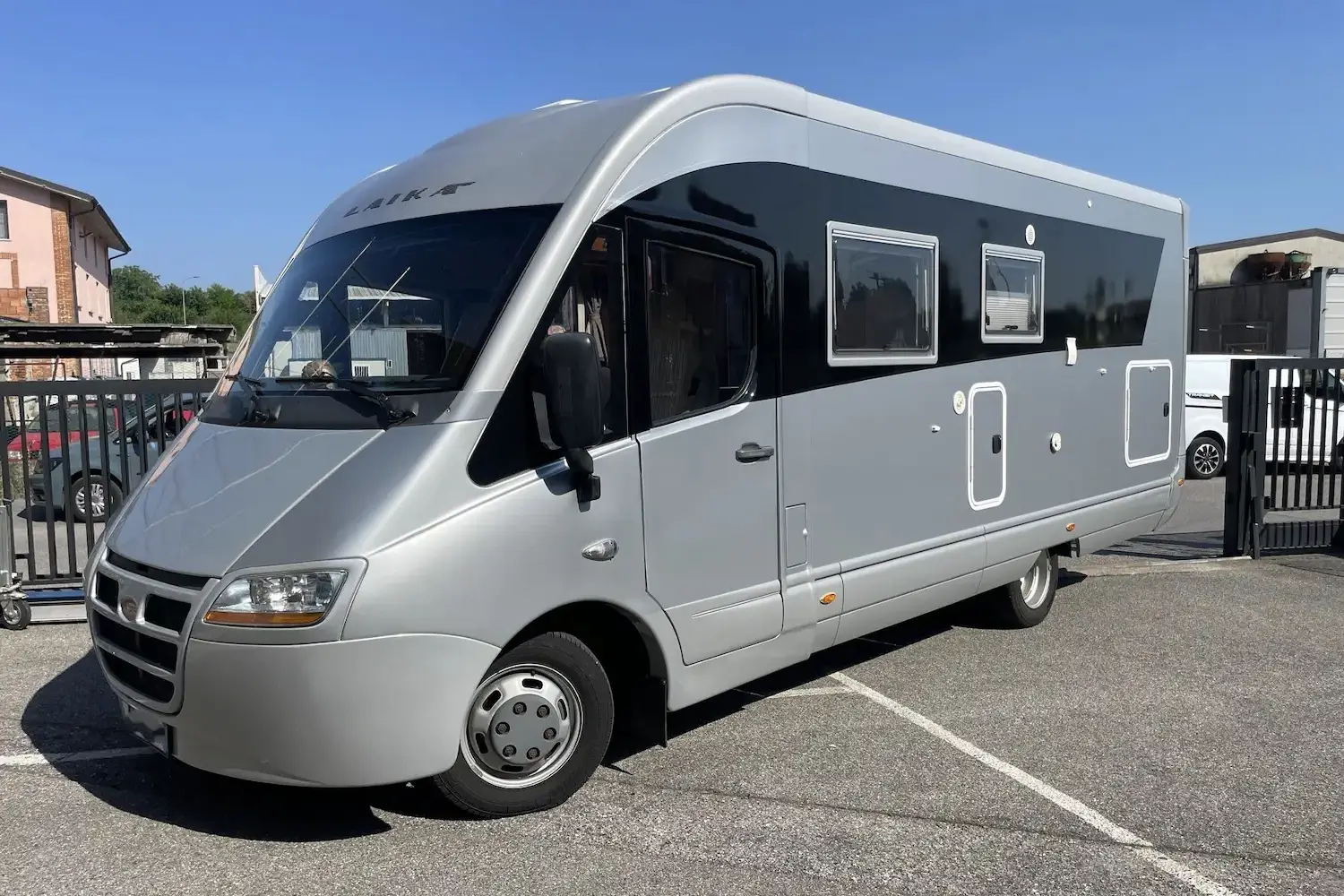Camper integrato Laika carrozzeria grigio argento e nero con profili bianchi, parcheggiato in un piazzale sotto un cielo azzurro.
