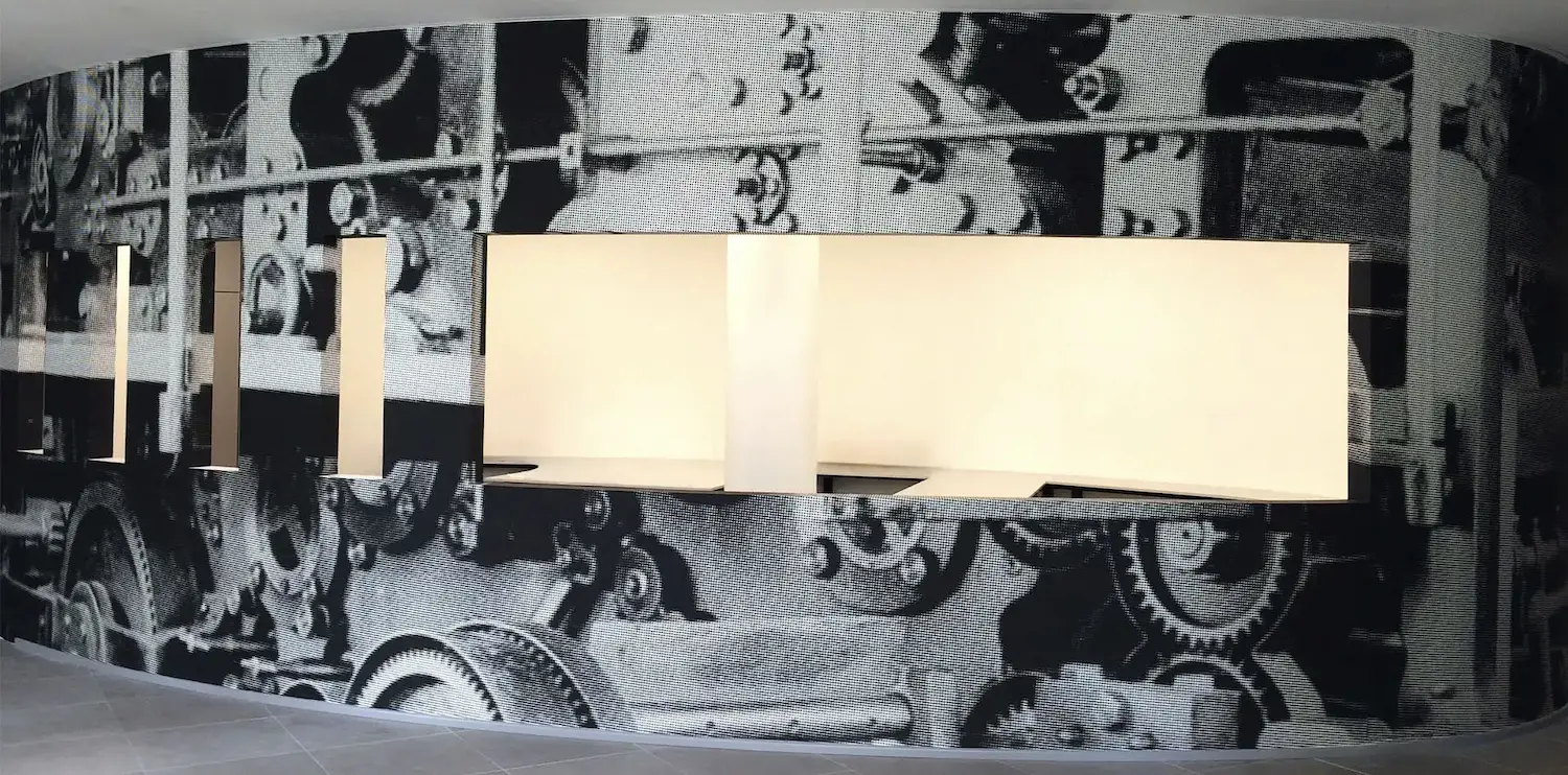 Parete curva rivestita da una stampa fotografica in bianco e nero raffigurante ingranaggi e macchinari industriali, con pannelli luminosi retroilluminati a incasso integrati nella parete.