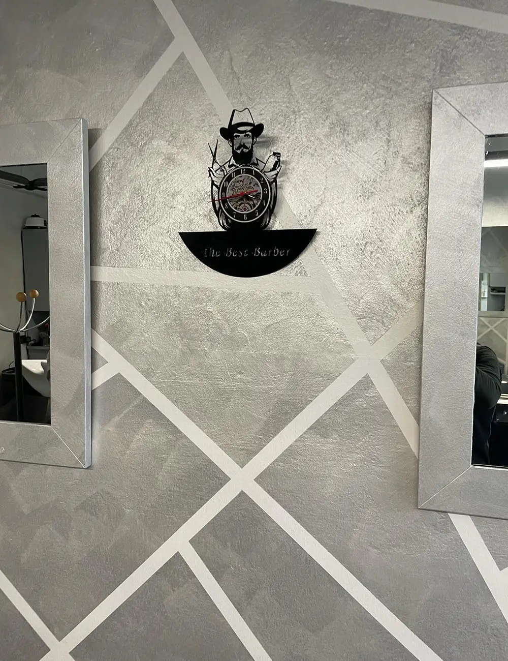 Parete decorativa di una barberia con rivestimento grigio effetto cemento, motivo geometrico a linee diagonali bianche, orologio decorativo a tema barber e specchi con cornice argentata.