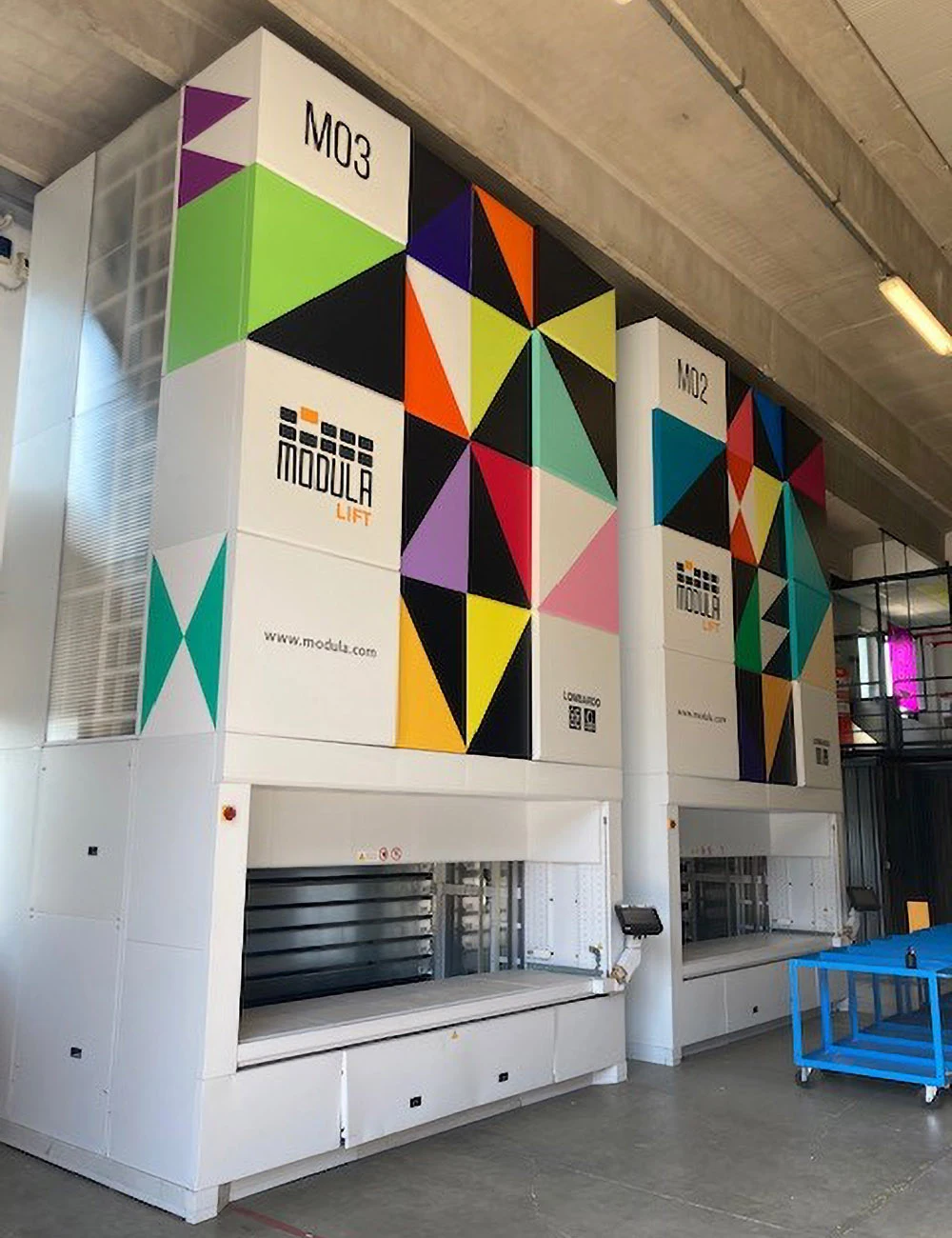 Due magazzini verticali automatici Modula Lift con pannellatura decorativa a motivi geometrici triangolari multicolore, installati all'interno di un capannone industriale.