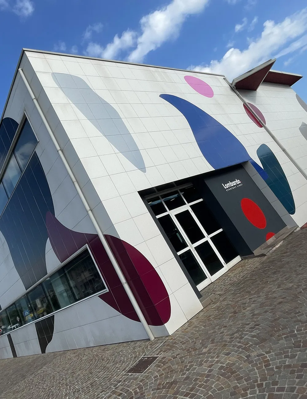 Facciata esterna della sede Lombardo Contract Store decorata con grandi forme geometriche astratte multicolore in blu, rosso, bordeaux e rosa su pannellatura bianca.