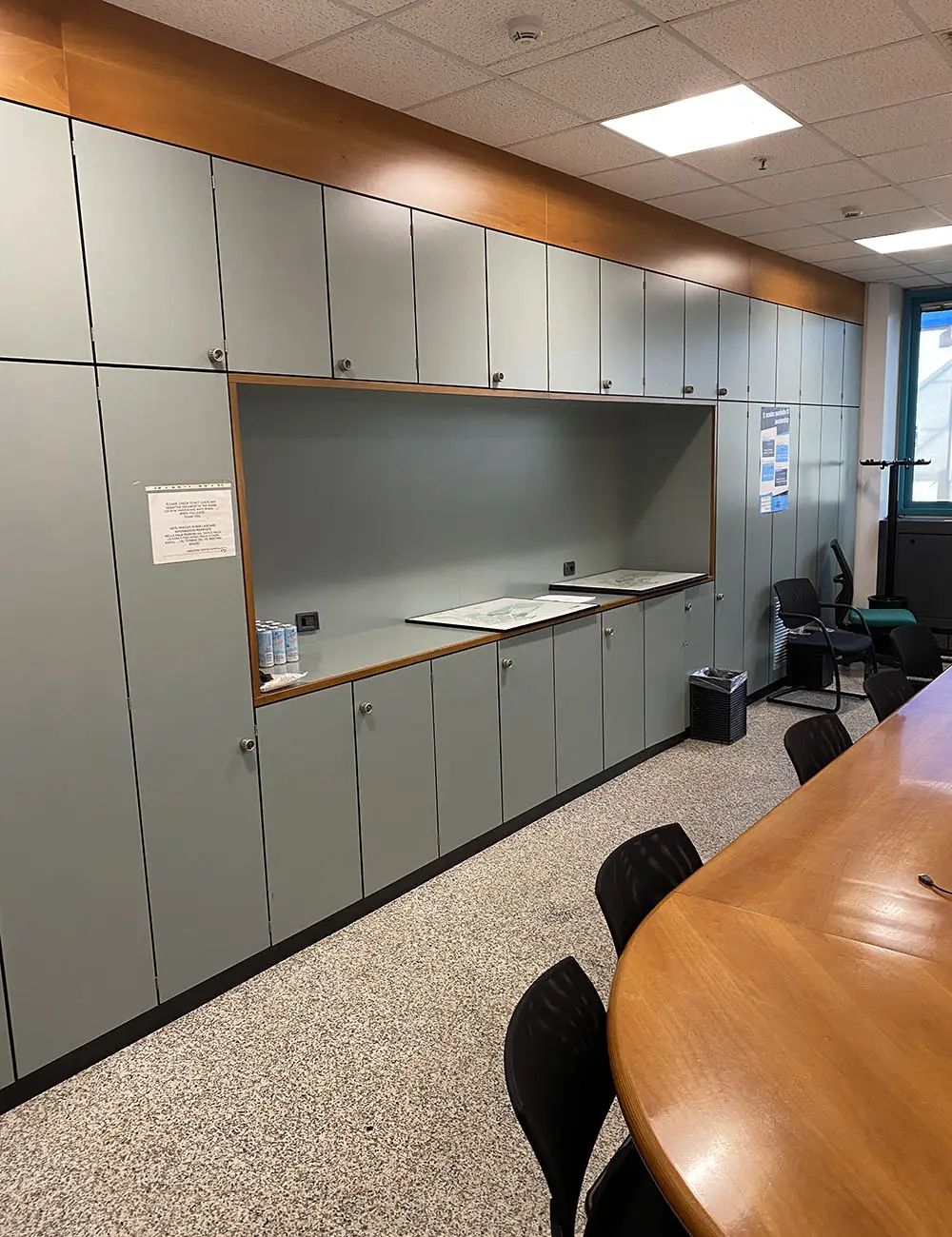 Sala riunioni con parete di armadi a tutta altezza rivestiti con pellicola grigio azzurro e profili in legno, tavolo ovale in legno, sedie nere e pavimento in moquette.
