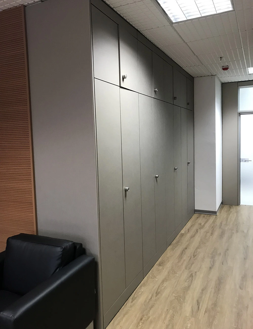 Corridoio di ufficio con parete di armadi a tutta altezza rivestiti con pellicola grigio opaco, maniglie in acciaio, pavimento in parquet chiaro e poltrona in pelle nera.