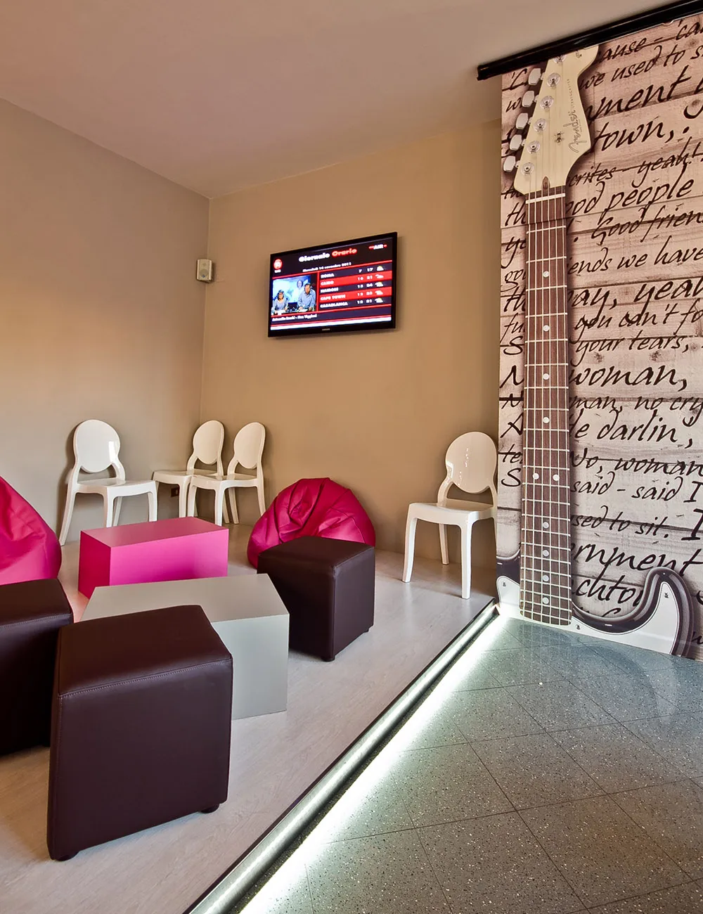 Sala d'attesa moderna con pouf e sedute in marrone e fucsia, televisore a parete e porta scorrevole rivestita con stampa fotografica raffigurante una chitarra elettrica Fender e testi musicali.
