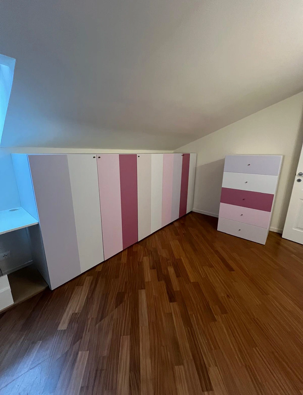 Camera con armadio a muro su misura e cassettiera rivestiti con pellicola a strisce verticali in tonalità bianco, rosa e ciclamino, pavimento in parquet e soffitto mansardato.