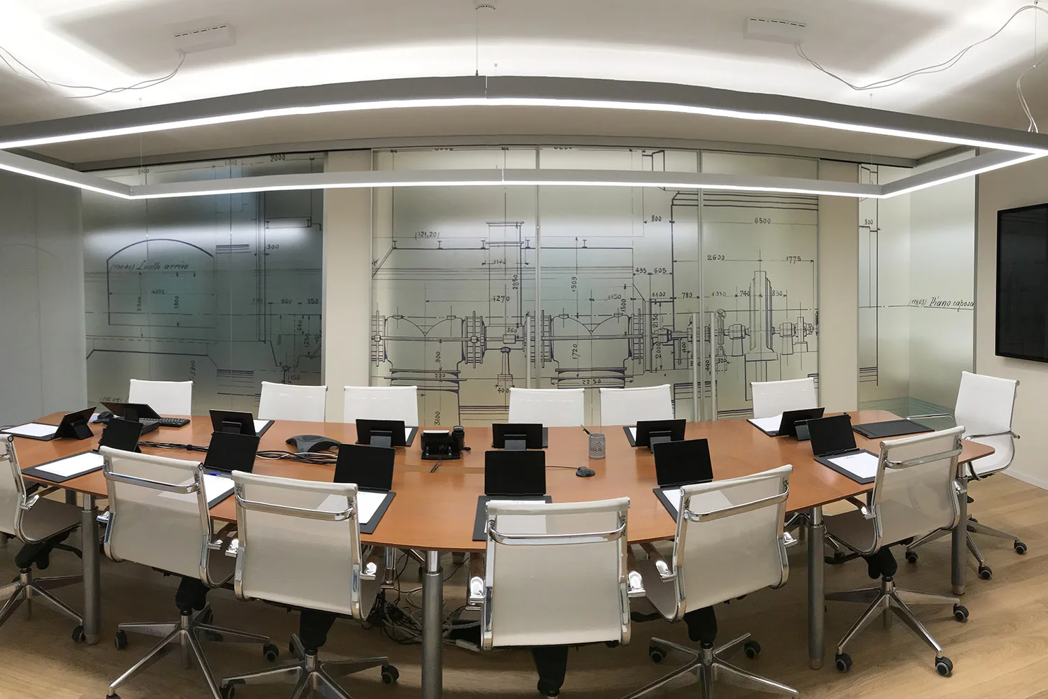 Sala riunioni elegante con tavolo ovale in legno, sedie bianche, illuminazione lineare curva a soffitto e parete in vetro decorata con stampa di un disegno tecnico industriale.