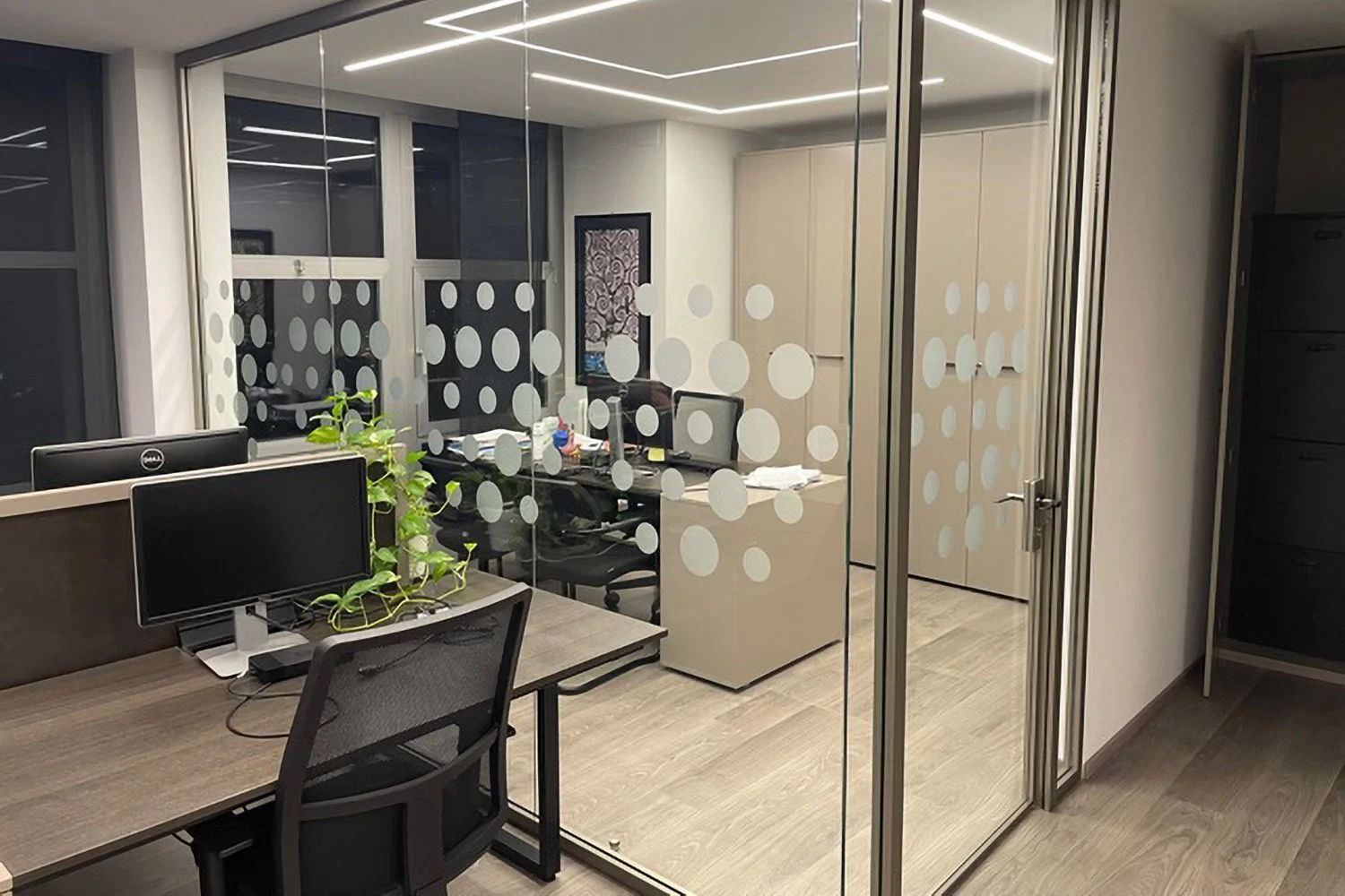 Ufficio privato con pareti divisorie in vetro decorate con cerchi bianchi satinati, postazione di lavoro con monitor, pianta decorativa, armadi beige e pavimento in parquet chiaro.