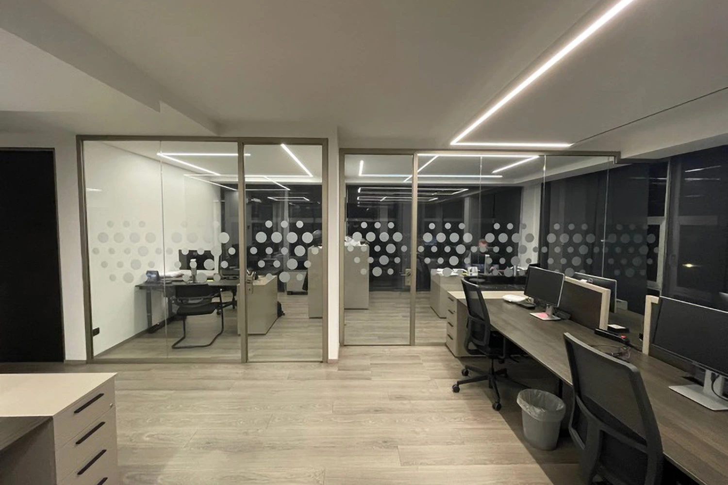 Spazio ufficio moderno con postazioni di lavoro, pareti divisorie in vetro decorate con cerchi bianchi satinati, illuminazione lineare a soffitto e pavimento in gres effetto legno.