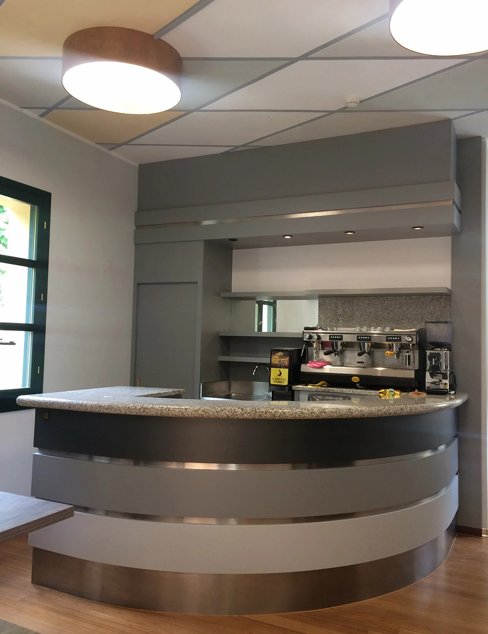 Interno di un bar moderno con bancone curvo rivestito in acciaio spazzolato e piano in granito, macchina per il caffè professionale e illuminazione a soffitto con lampade circolari in legno.