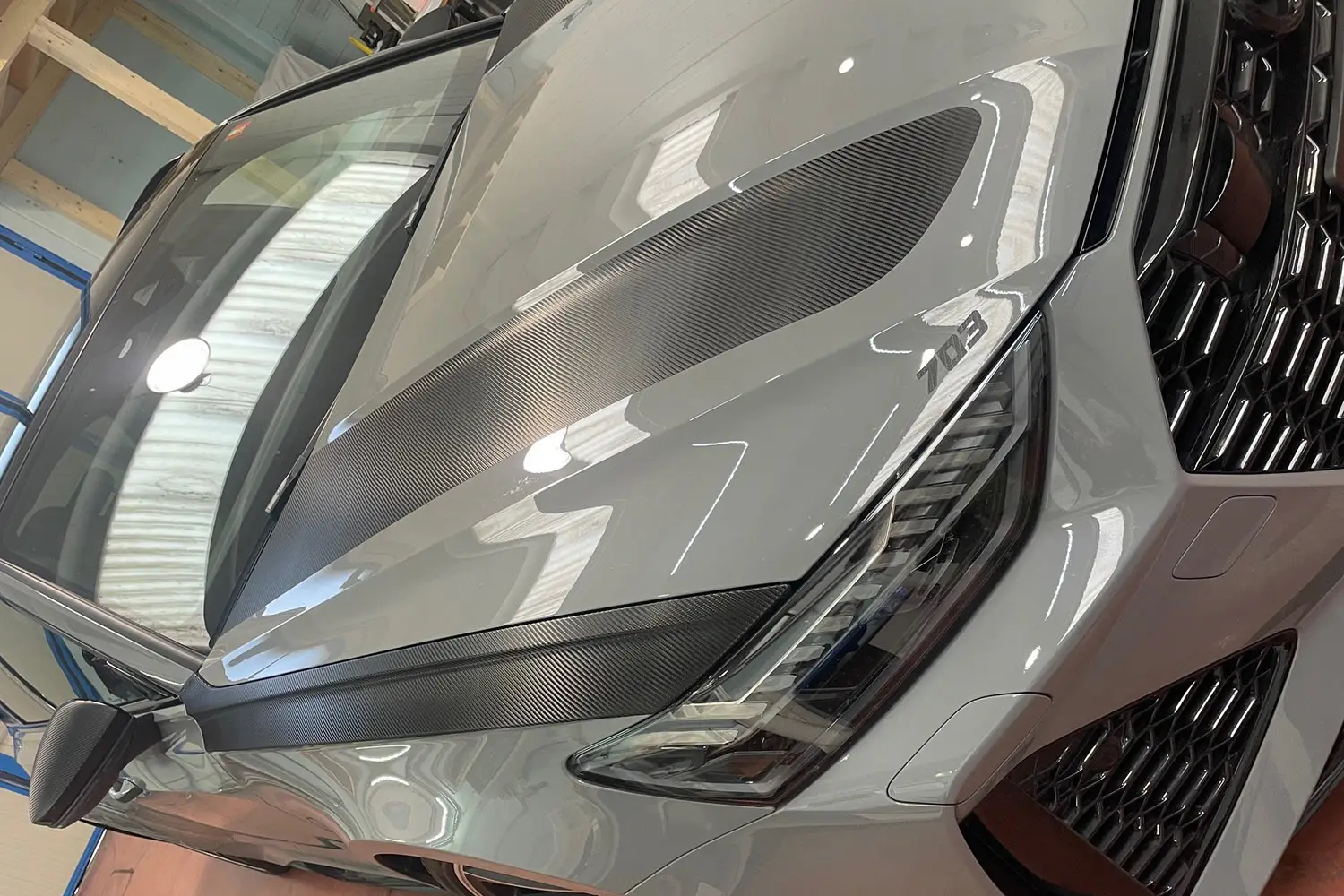 Dettaglio del cofano anteriore di una Audi RS6 grigio metallizzato con inserti decorativi in pellicola effetto carbonio applicati sul cofano e sui contorni dei fari.